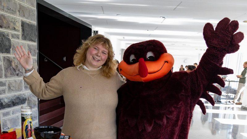 The HokieBird mascot and a blond woman in a beige sweater wave viewers.