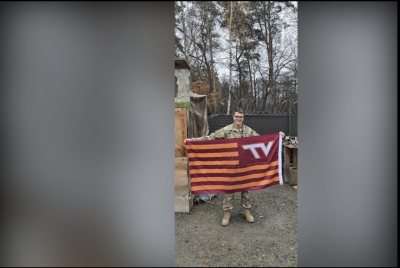 A serviceman in a camouflage uniform stands in front of barbed wire and chain link fencing holding a Virginia Tech flag. 